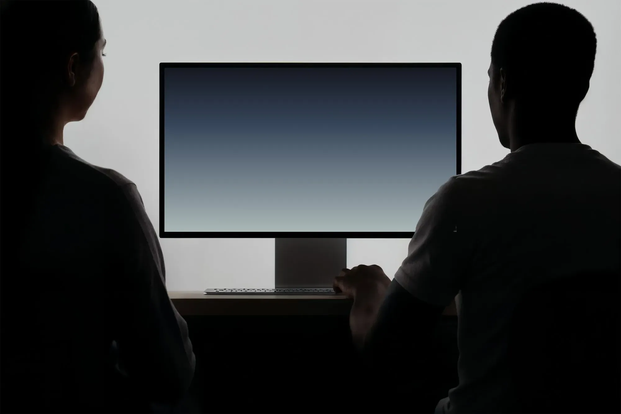 Siluety dvou lidí před velkým monitorem s modrým přechodovým obrazem.
